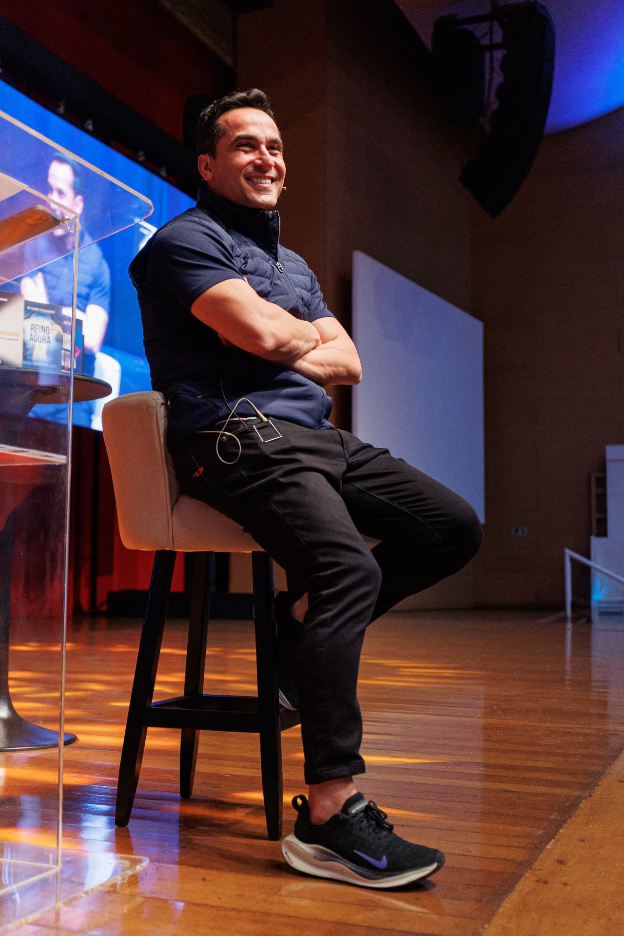 Márcio Micheli sentado num banquinho no palco, de braços cruzados, sorrindo